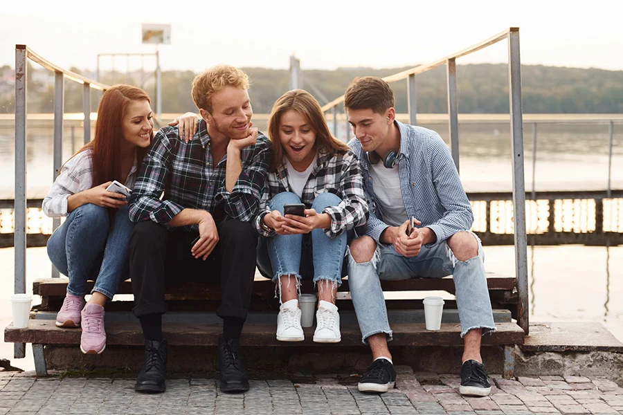 Four Smiling Friends Sitting Together Outdoors And Looking At A Phone, Symbolizing Social Connection In Bed Recovery. four smiling friends sitting together outdoors and looking at a phone, symbolizing social connection in bed recovery.