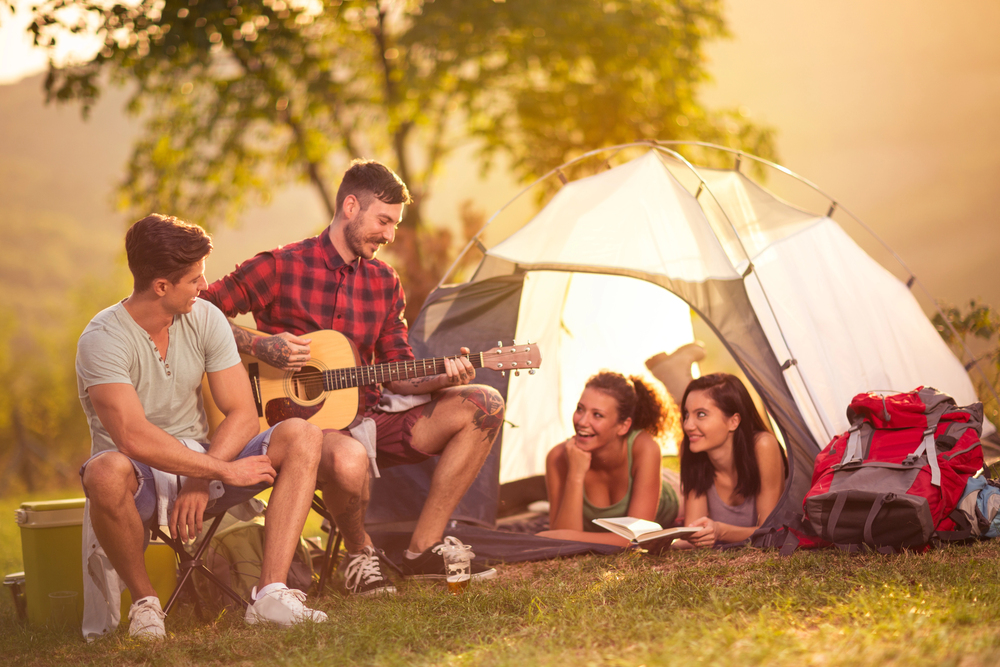 a group of friends relax at a campsite at sunset, with one playing guitar while the others lounge and laugh beside a tent.
