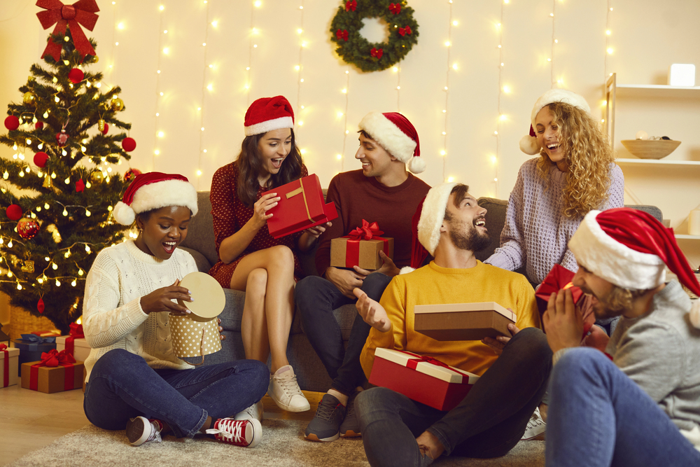 The Ultimate Guide To Navigating Holidays With An Eating Disorder 3 friends in santa hats exchange gifts by a glowing christmas tree, laughing together in a cozy, festive living room.