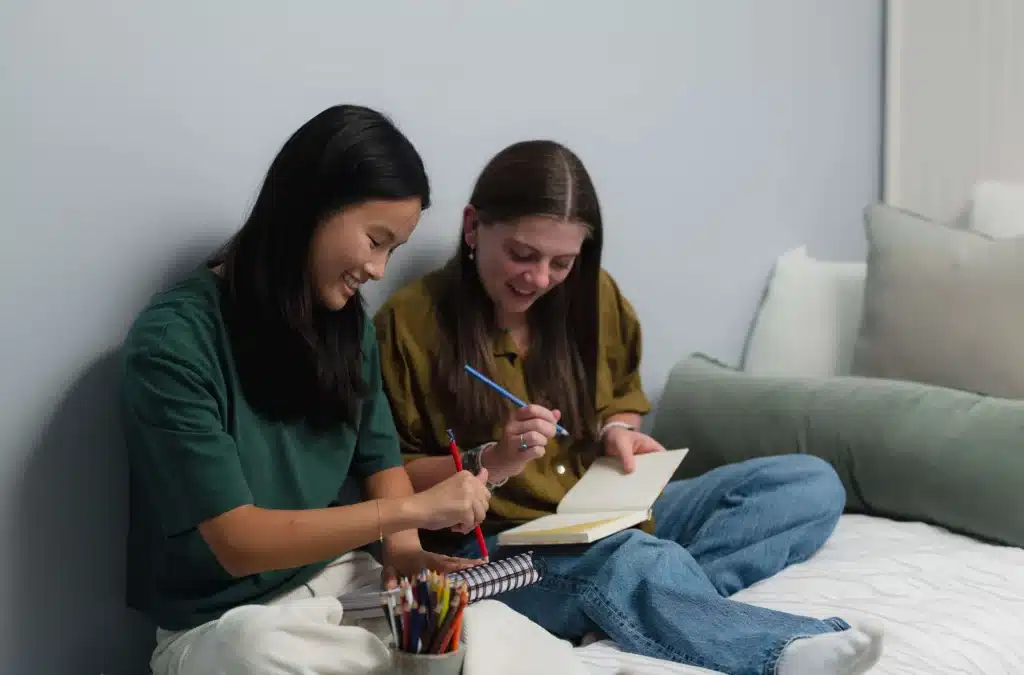 two young women sitting together on a bed, smiling and writing in journals with colored pencils, representing compassionate support for eating disorders at newcircle.