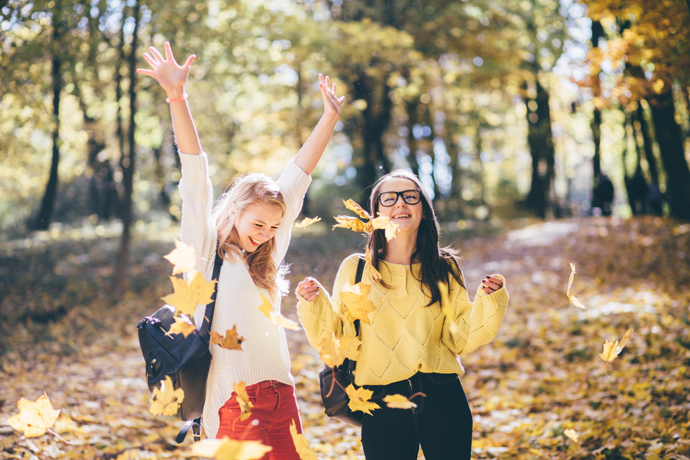 two teenage friends laugh and toss colorful autumn leaves in a sunlit park, wearing cozy sweaters and backpacks.